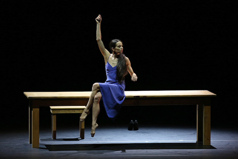 Vishenva in a blue camisole dress sits on the long wooden table with her legs crossed